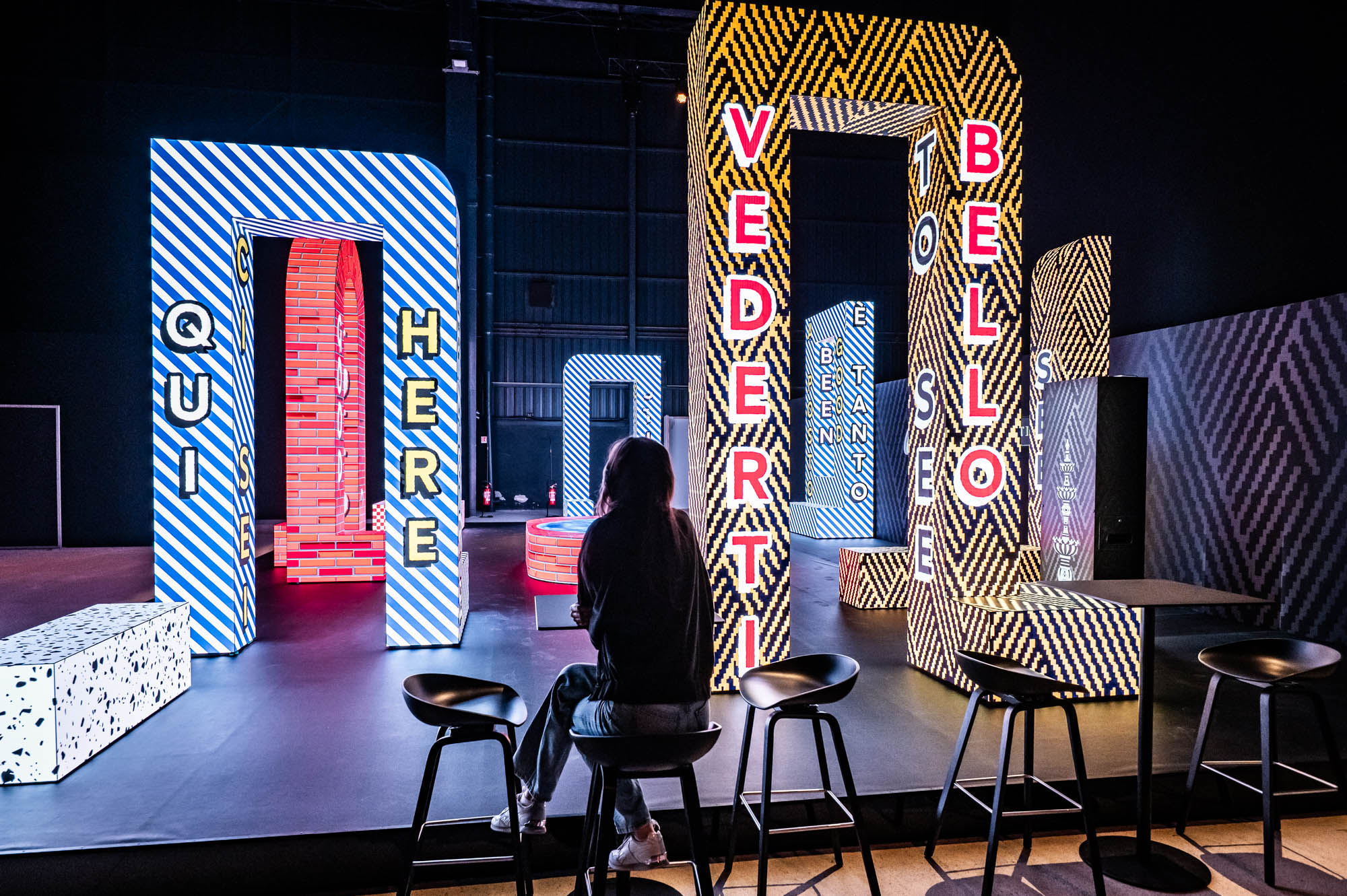 Woman seated at a bar in front of tall illuminated letter sculptures with bold graphic patterns.