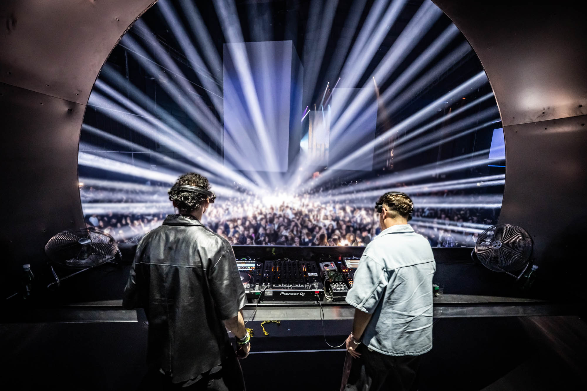 DJs performing on stage, facing the crowd under radiating light beams.
