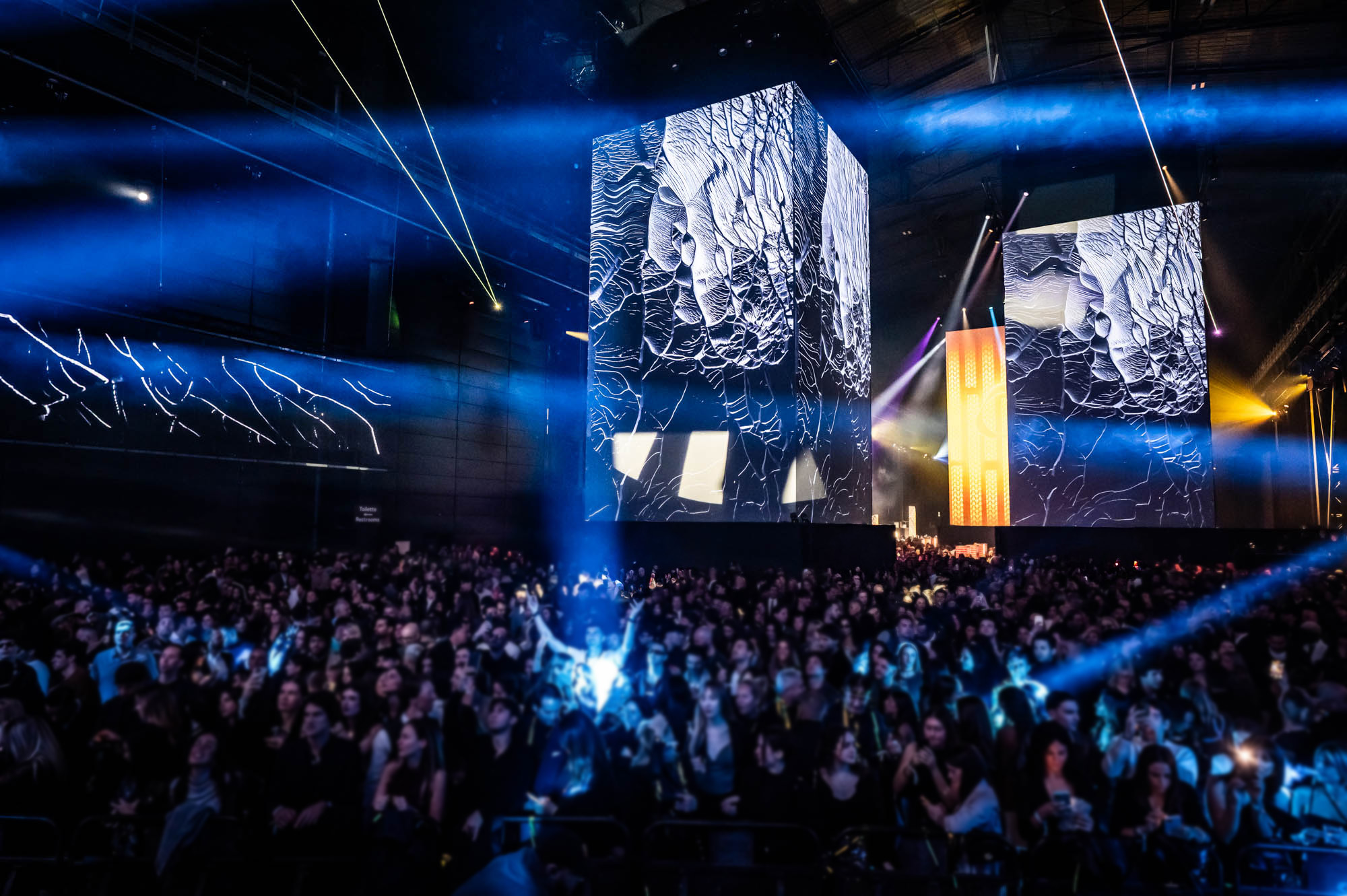 Crowd under blue light beams in front of projection-mapped towers at Sensorium Worlds.