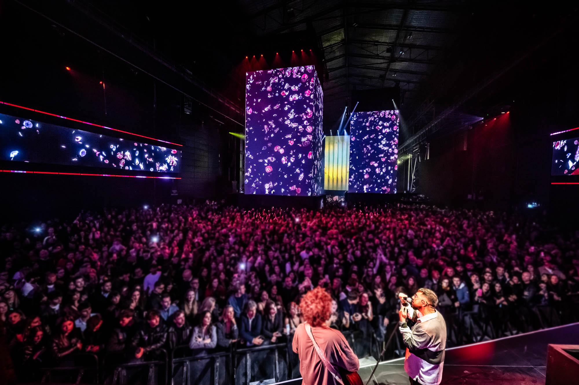 Wide view of a large crowd in red light facing towers with purple visuals.