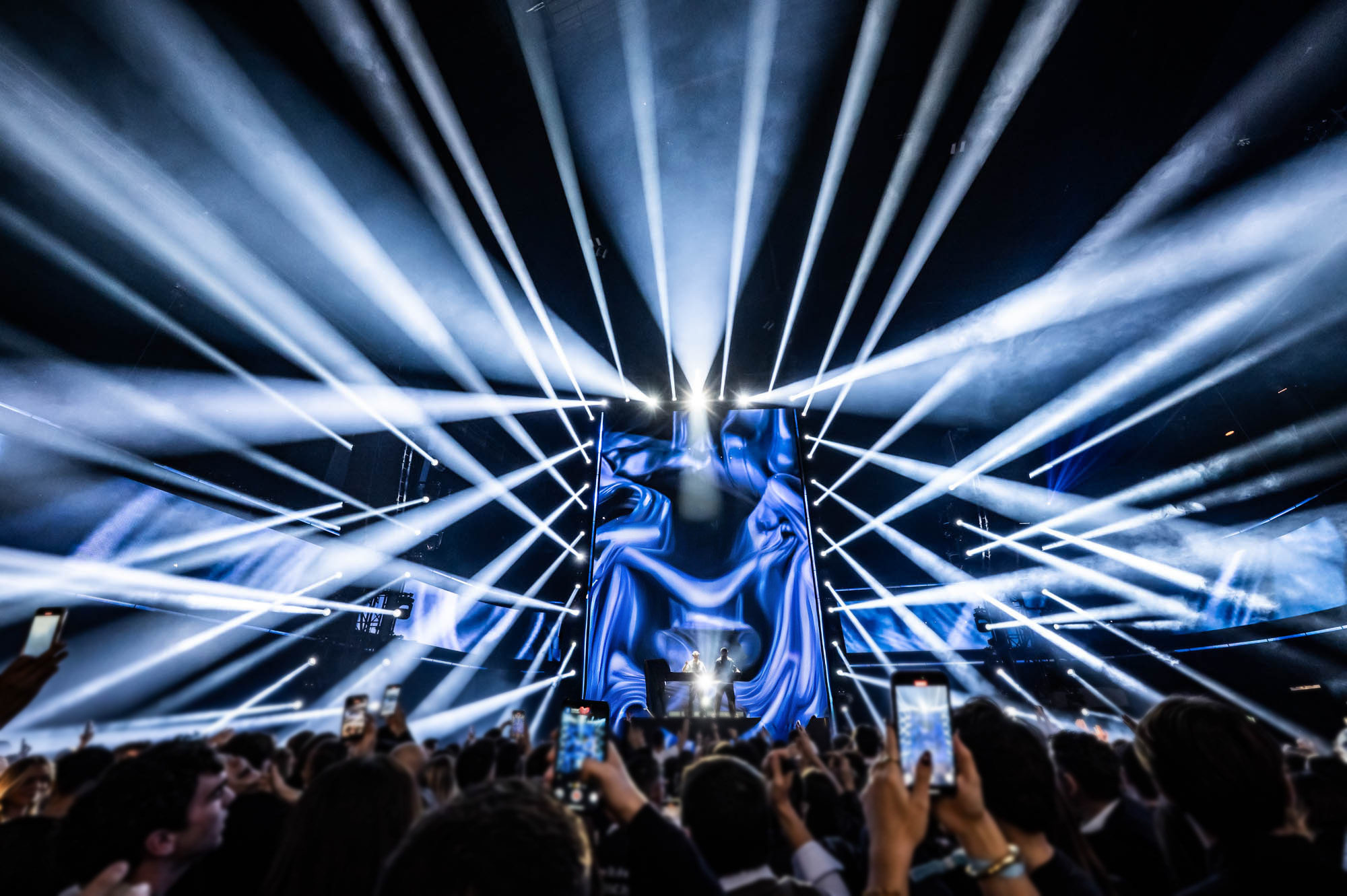 Headliner on stage under radiating white light beams, with guests filming on their phones.