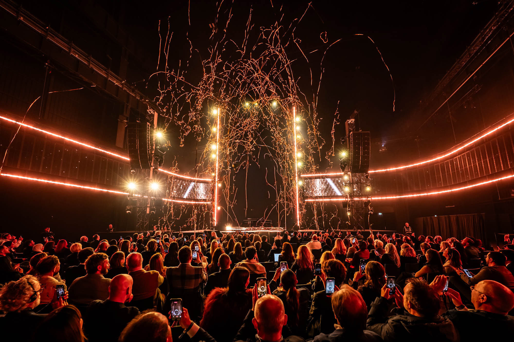 Seated audience facing a stage with orange light lines and falling confetti.