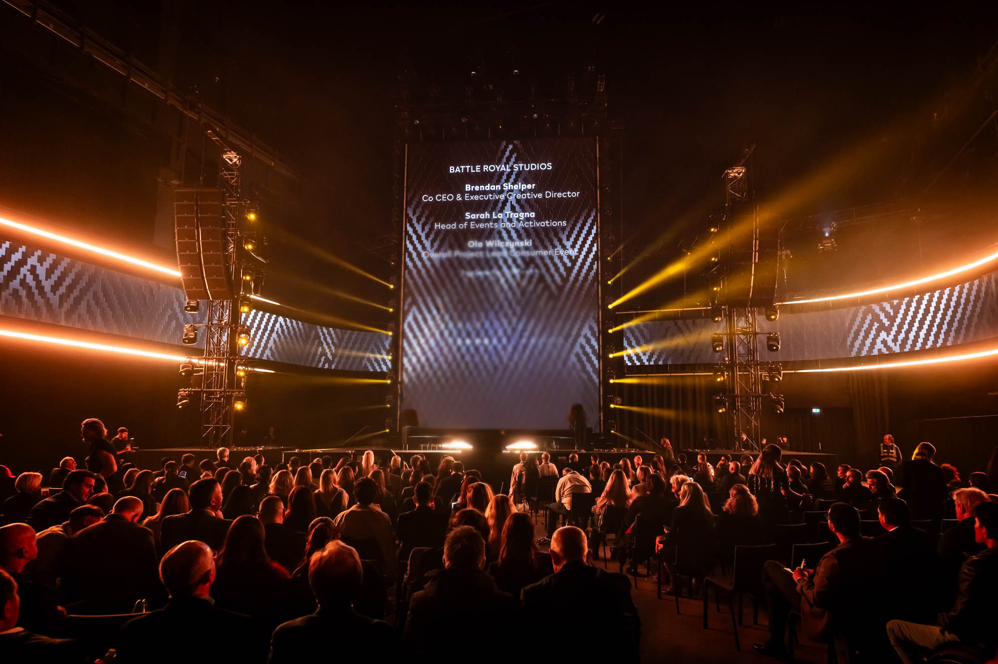 Press preview audience facing a stage framed by warm light beams and a central LED screen.