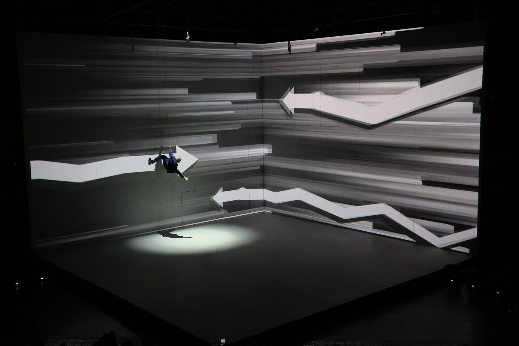 A performer hangs on a dark corner wall surrounded by projected white arrows and jagged lines, lit from below by a spotlight.
