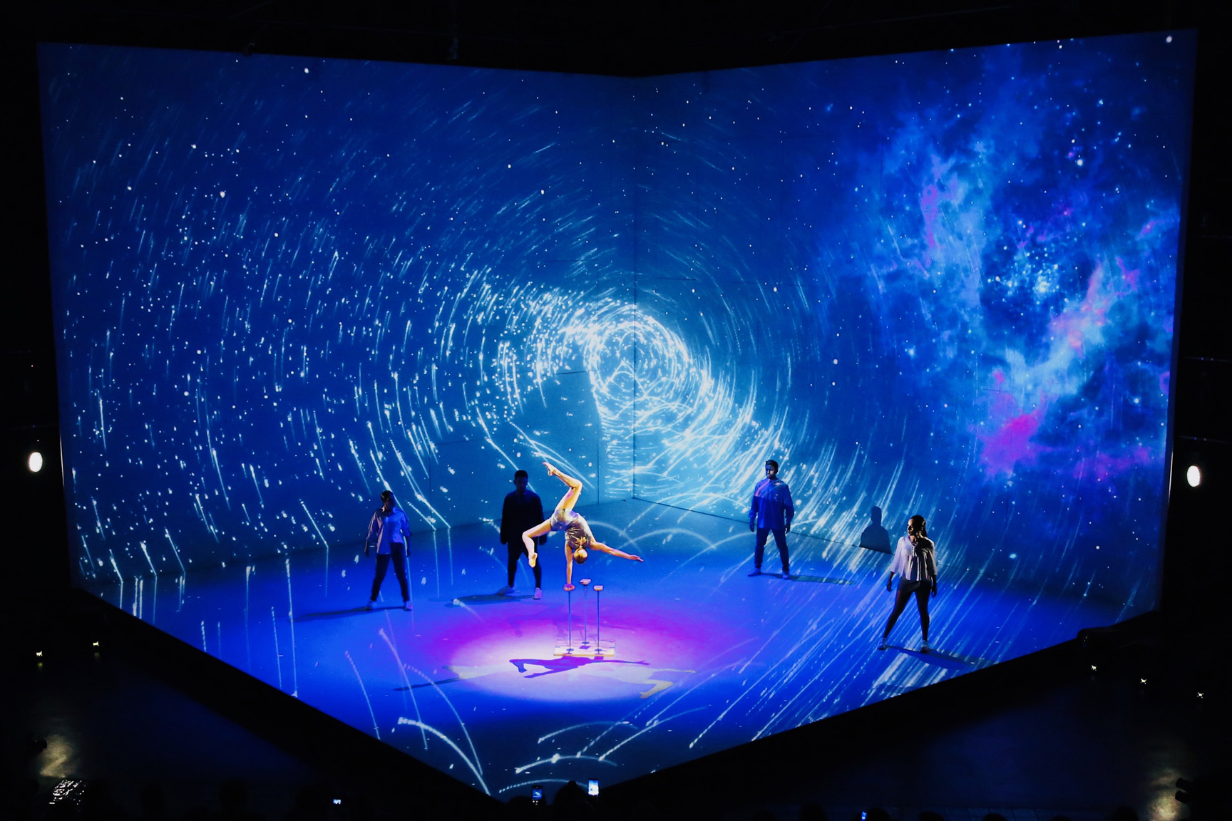 Two acrobats perform a lift in a pool of purple light while dancers stand around them, in front of a corner wall projected with a blue galaxy tunnel.