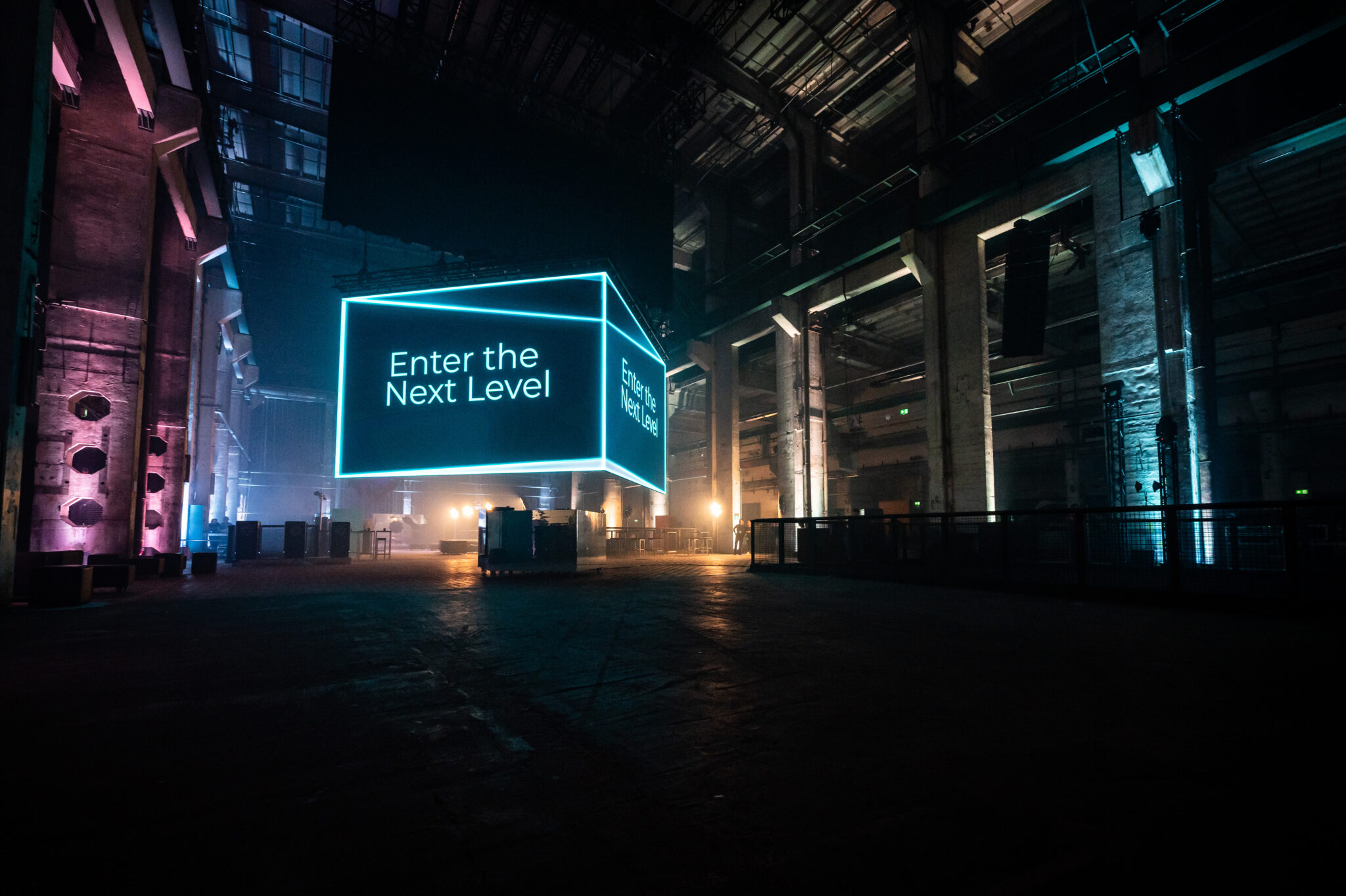 A glowing cube with white text floats above the floor of a long dark industrial hall, with pink and teal lights along the walls.