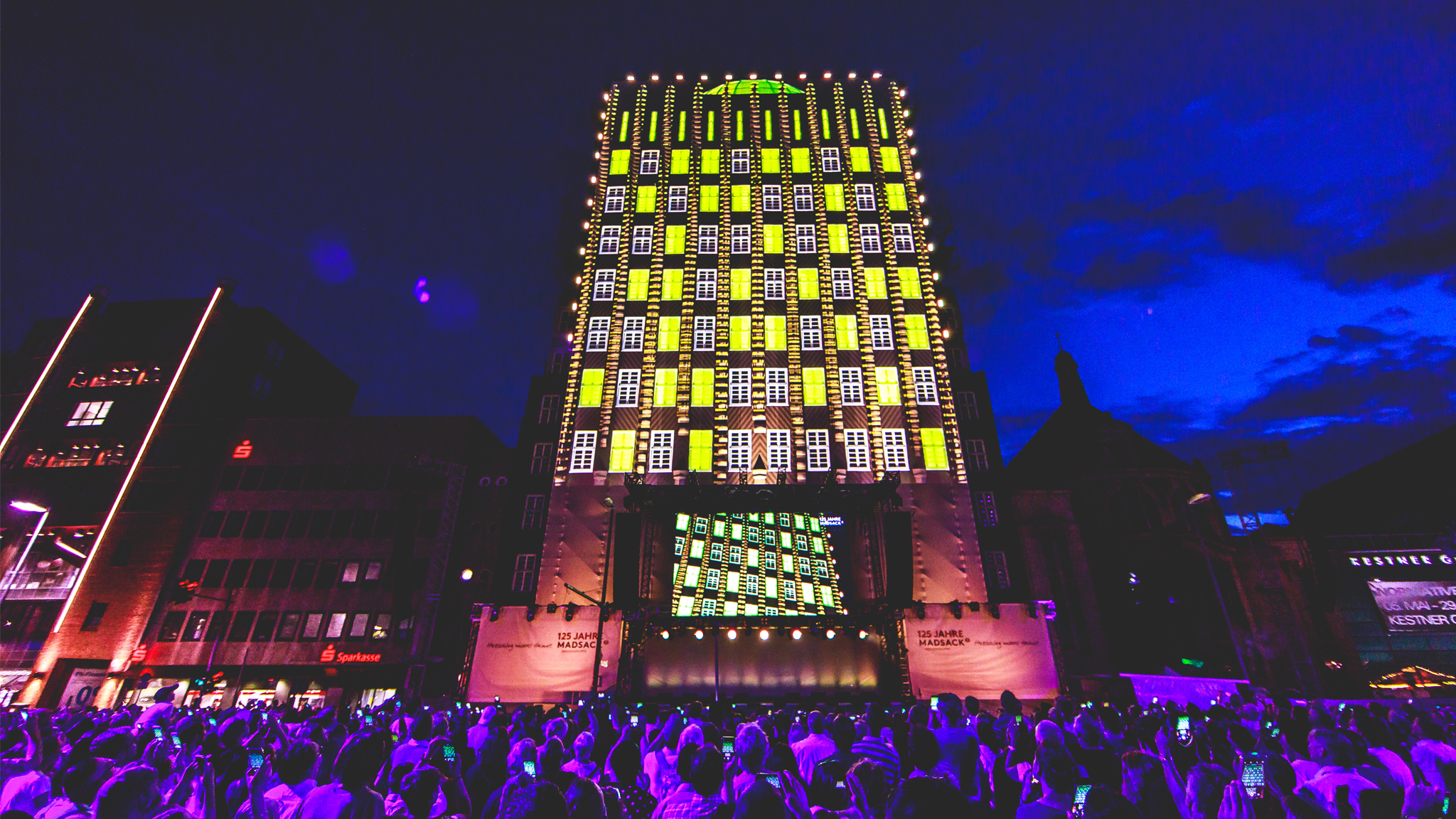 A tall building at dusk with its windows projected as a grid of glowing yellow and green panels, above a crowd lit in purple.