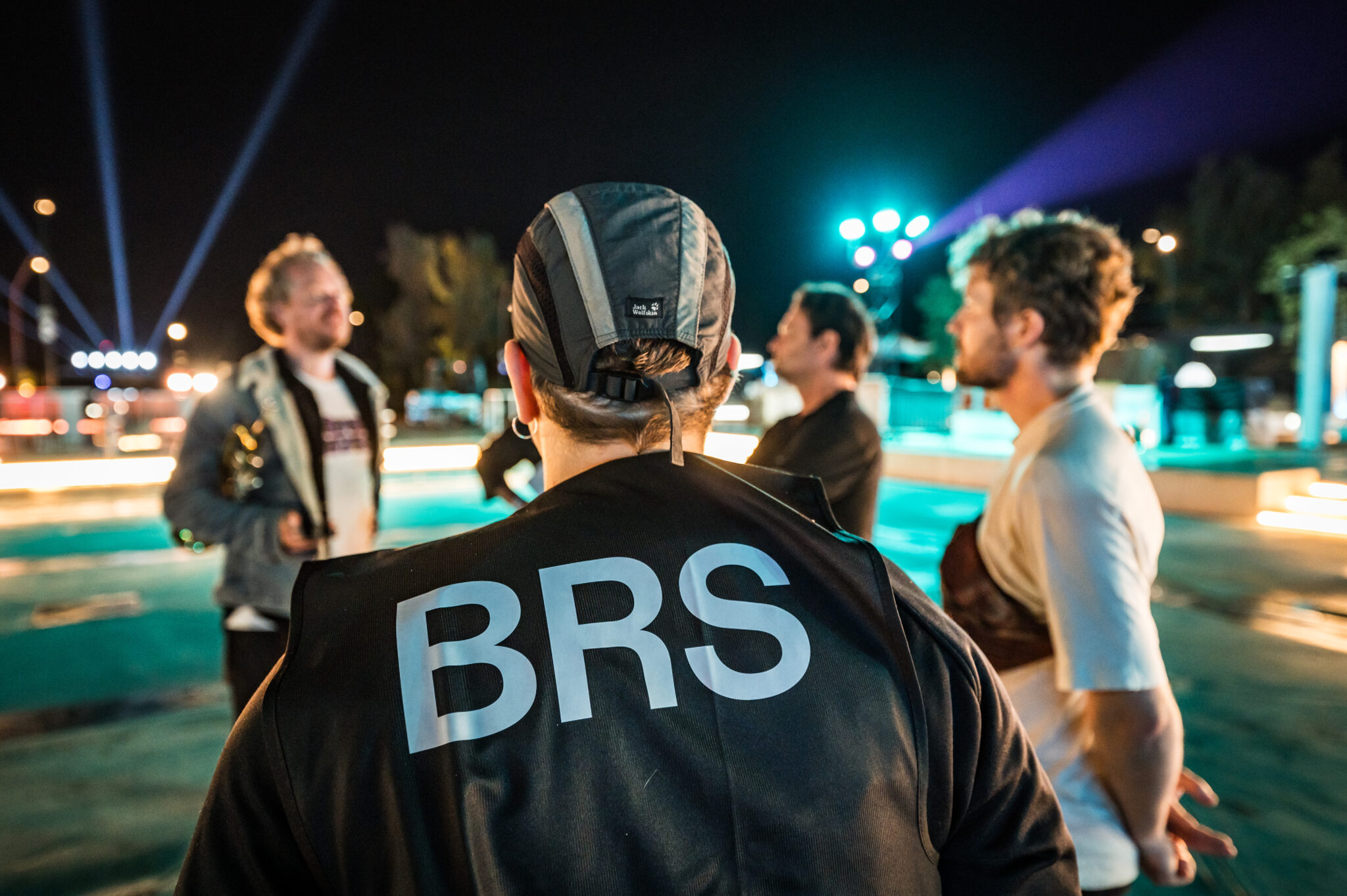A crew member in a cap and black jacket, seen from behind, talks with two guests at a night event lit by blue and teal searchlights.