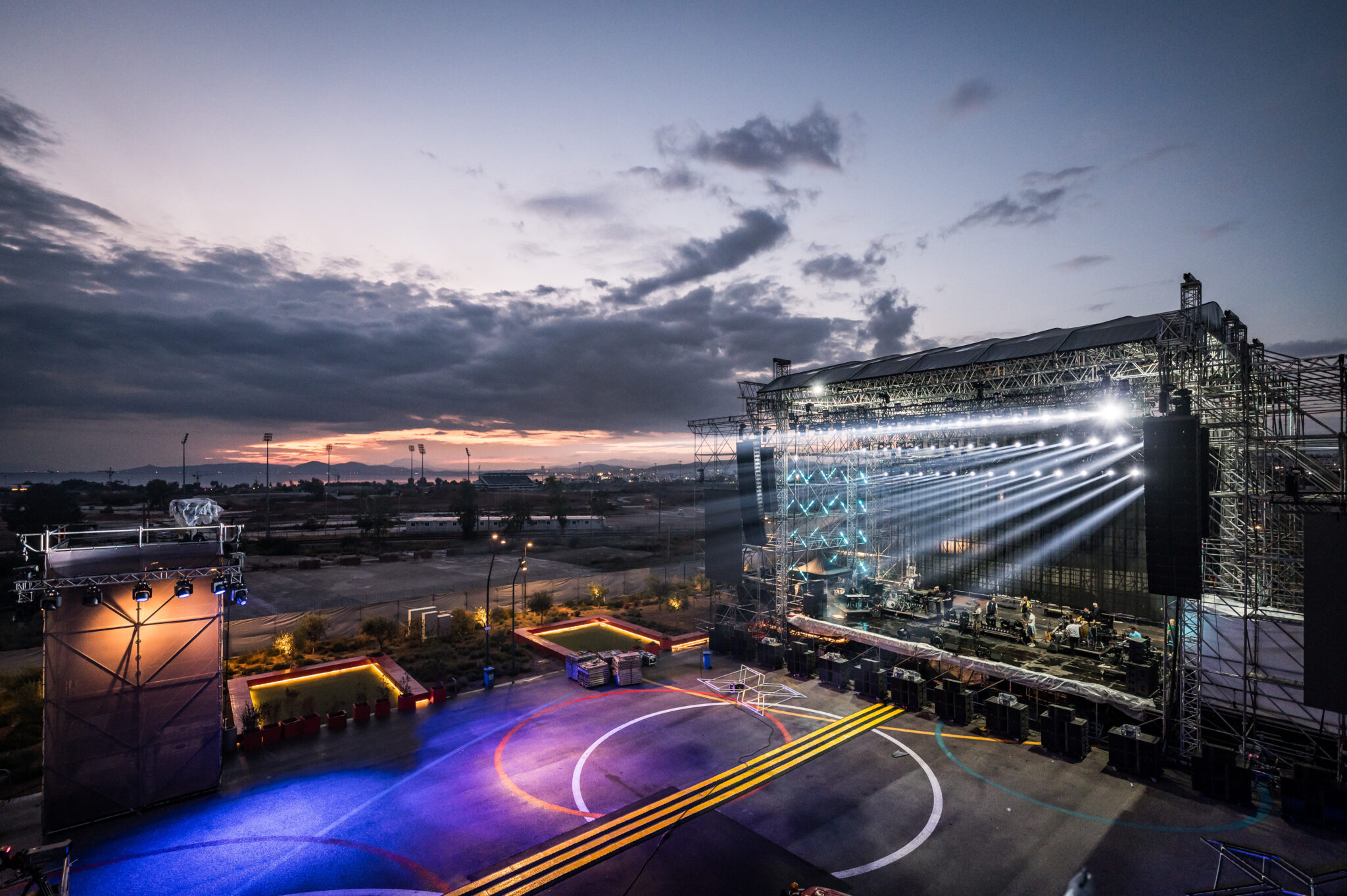 Aerial view at dusk of an open-air event site with a large lit stage, light beams and glowing floor graphics under a cloudy sky.