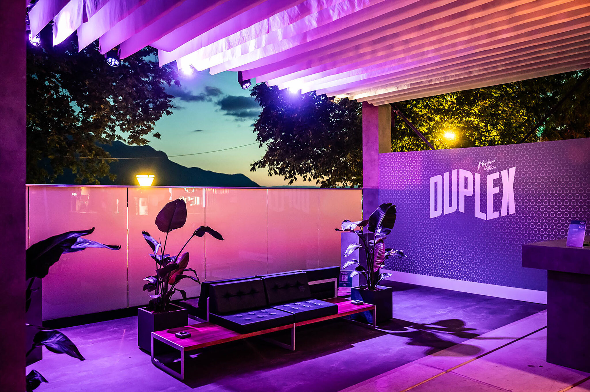 Terrace lounge at dusk with a Duplex logo wall, plants and a view of the lake and mountains.