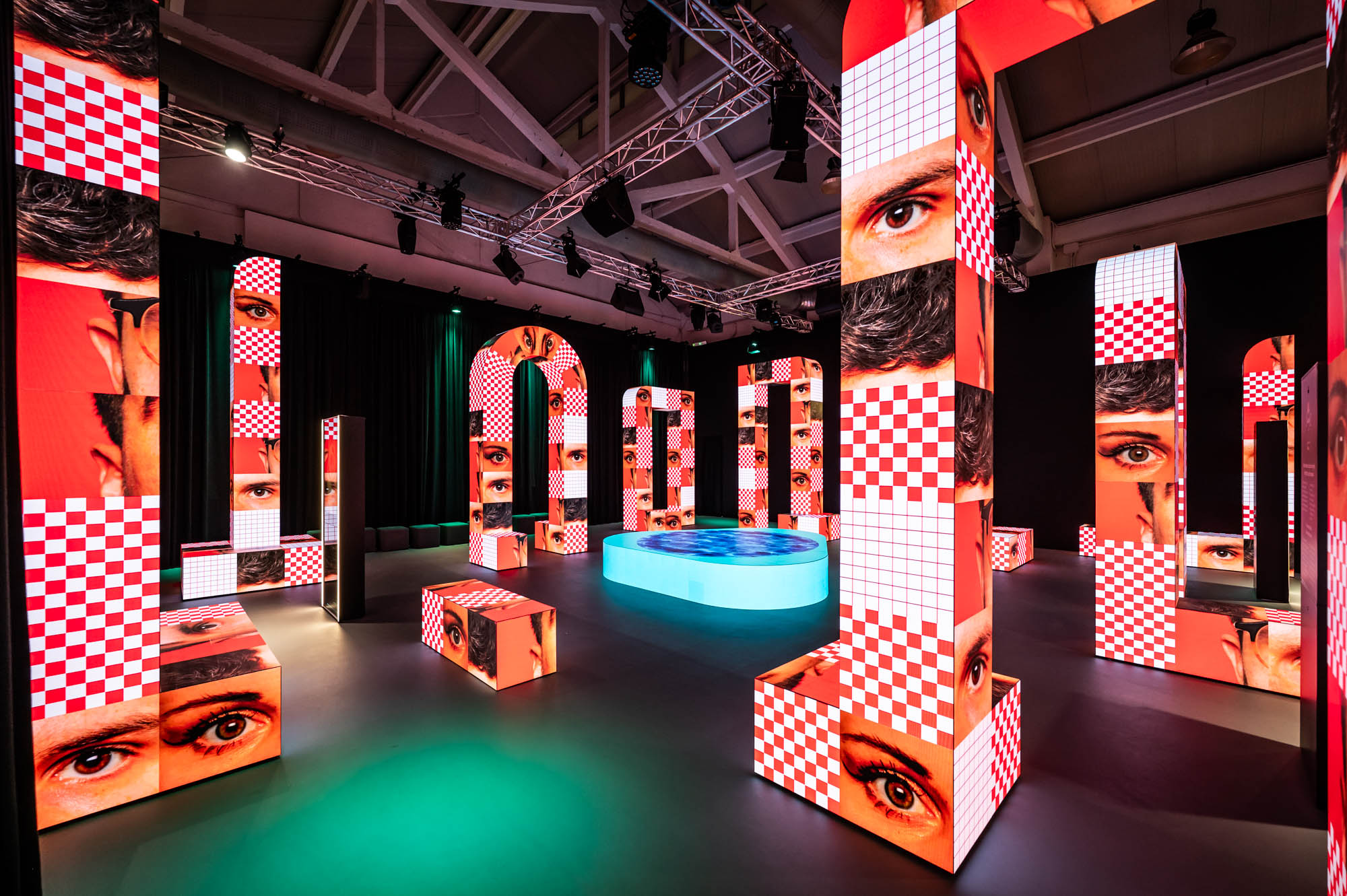 Dark hall filled with tall illuminated columns and arches showing close-ups of eyes and red-and-white checkerboard patterns, around a glowing turquoise oval pool.