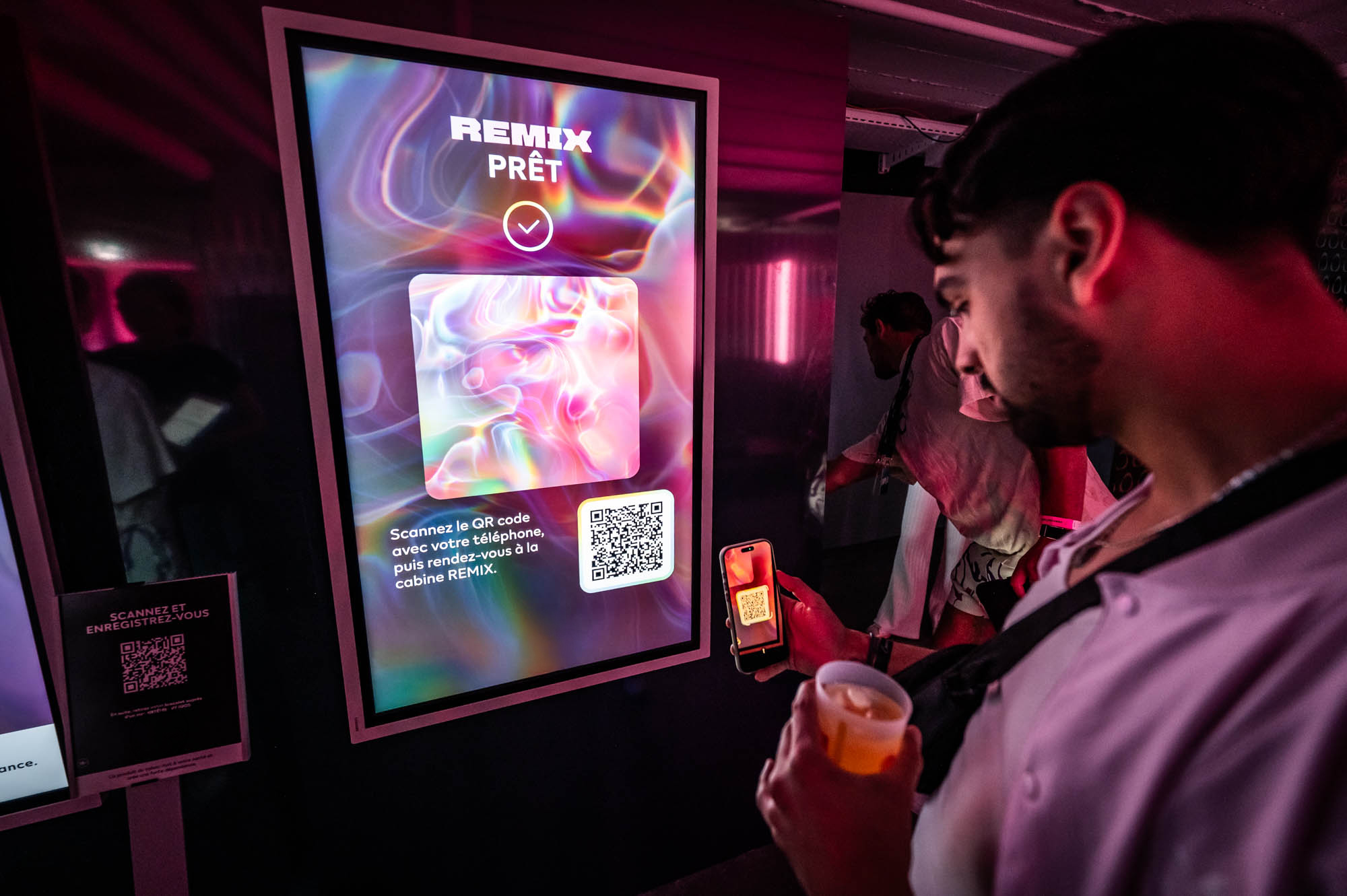 Guest holding a drink and phone in front of a Remix prêt screen displaying artwork and a QR code.