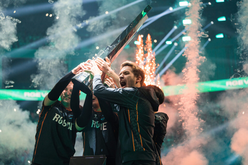 Players hold up a trophy amid confetti and smoke on a brightly lit stage.